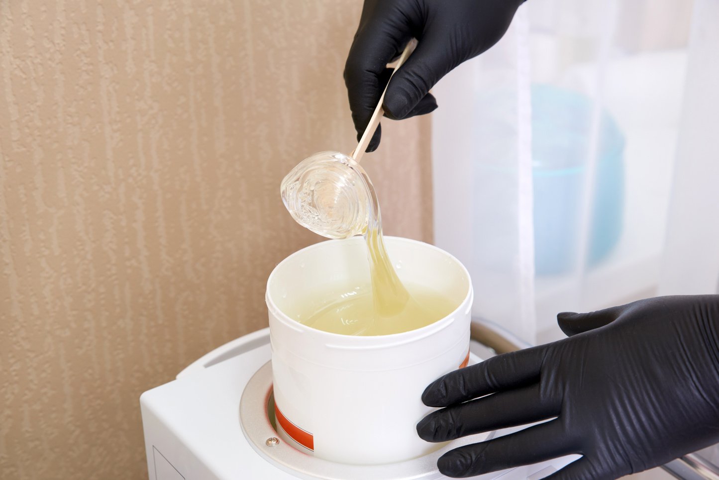 Wax heater with hot yellow wax on wooden spatula in human hand. Hot wax for depilation. Salon equipment